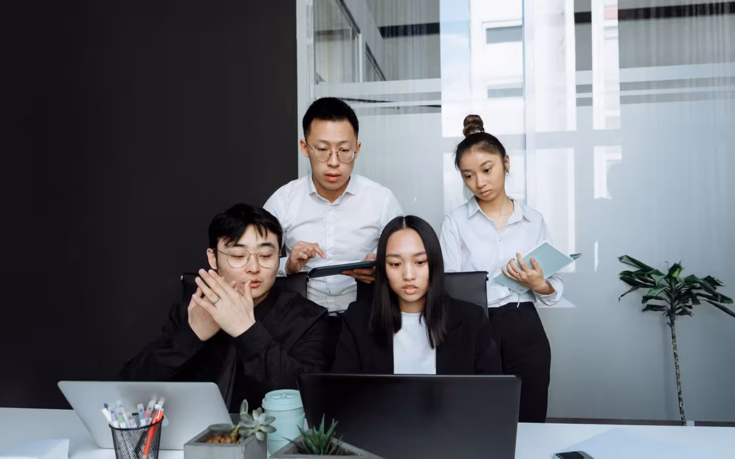 Four office workers gather around laptops and a tablet in a modern workspace.