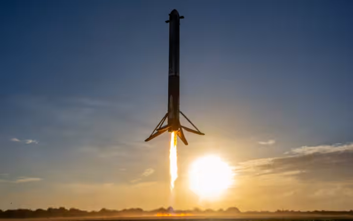 A SpaceX rocket hovers above a field, firing its engines at sunset.