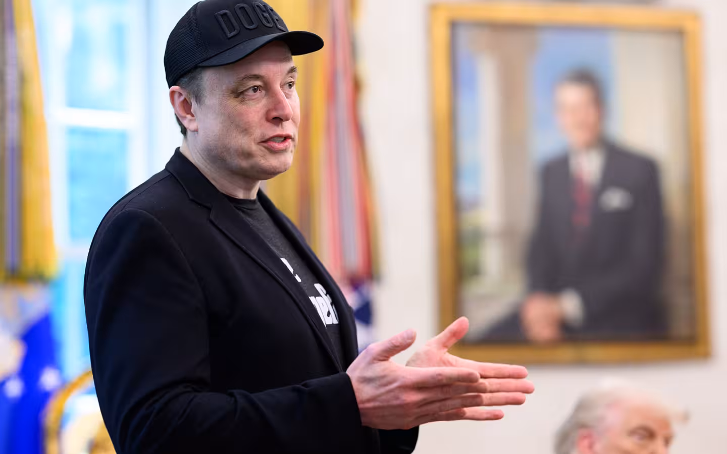 Elon Musk gestures while speaking in the Oval Office, with Donald Trump partly visible.