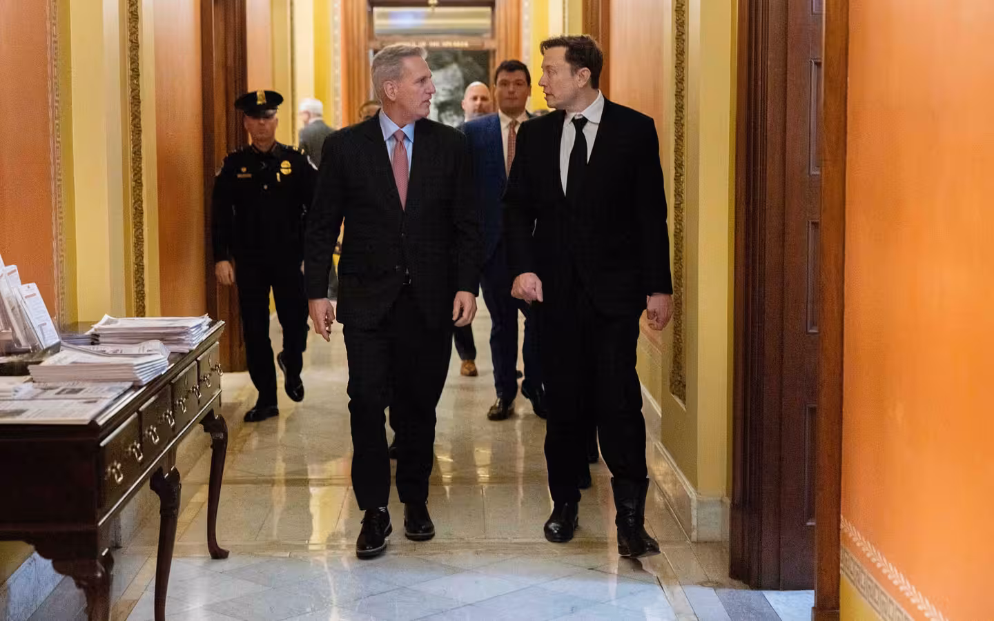 Two men in dark suits walking together down a congressional hallway