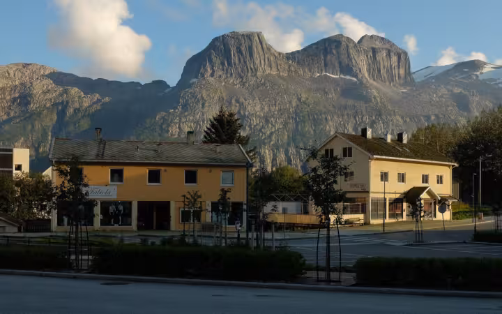 Buildings and a street in Glomfjord beneath steep, sunlit mountains