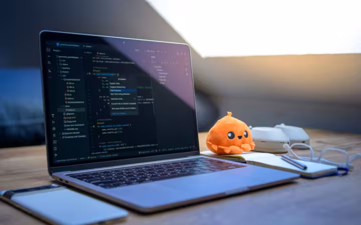 A cozy workspace featuring a laptop displaying code, a smartphone, and a cute orange plush toy on the desk.