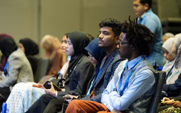 Conference attendees seated in rows, listening in an indoor hall.