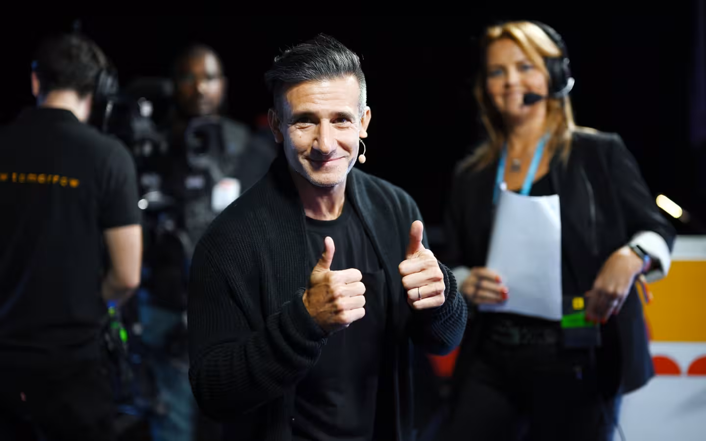 Josh Wolfe gives two thumbs-up beside camera crew and a headset-wearing presenter.