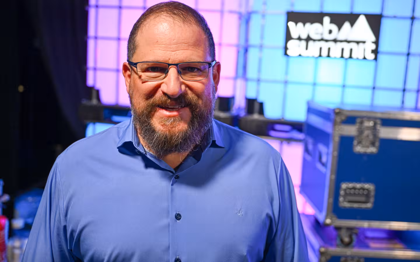Cristiano Amon smiles backstage at Web Summit amid colorful lighting and equipment.