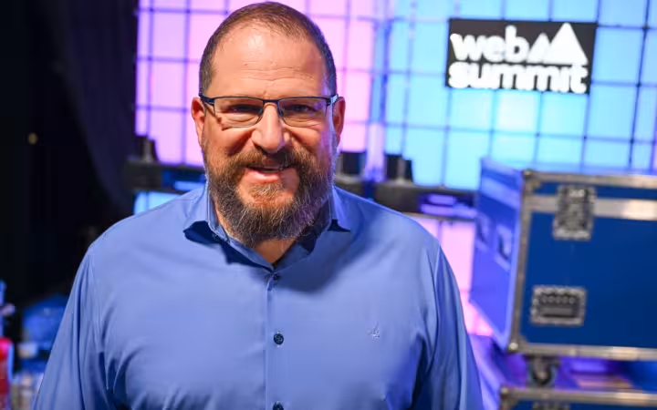 Cristiano Amon smiles backstage at Web Summit amid colorful lighting and equipment.
