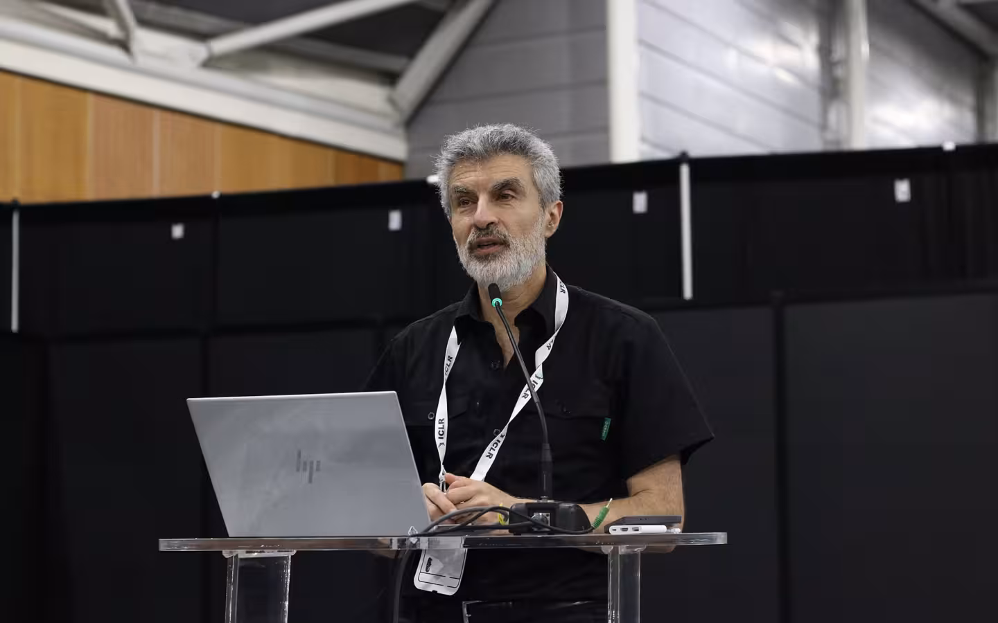 Yoshua Bengio speaks into a microphone behind a laptop at ICLR 2025.
