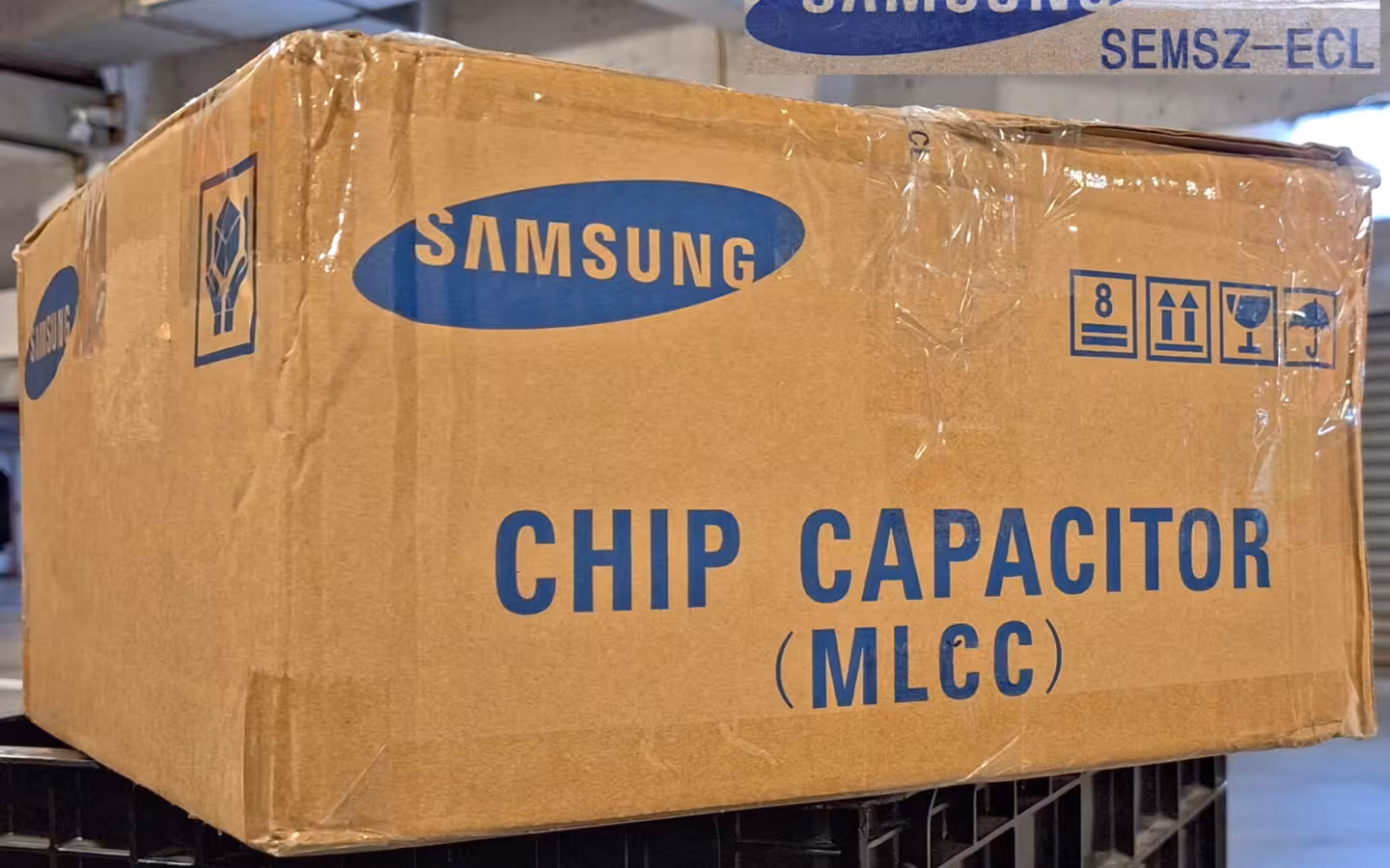 A Samsung Electro-Mechanics capacitor carton stacked on black crates indoors