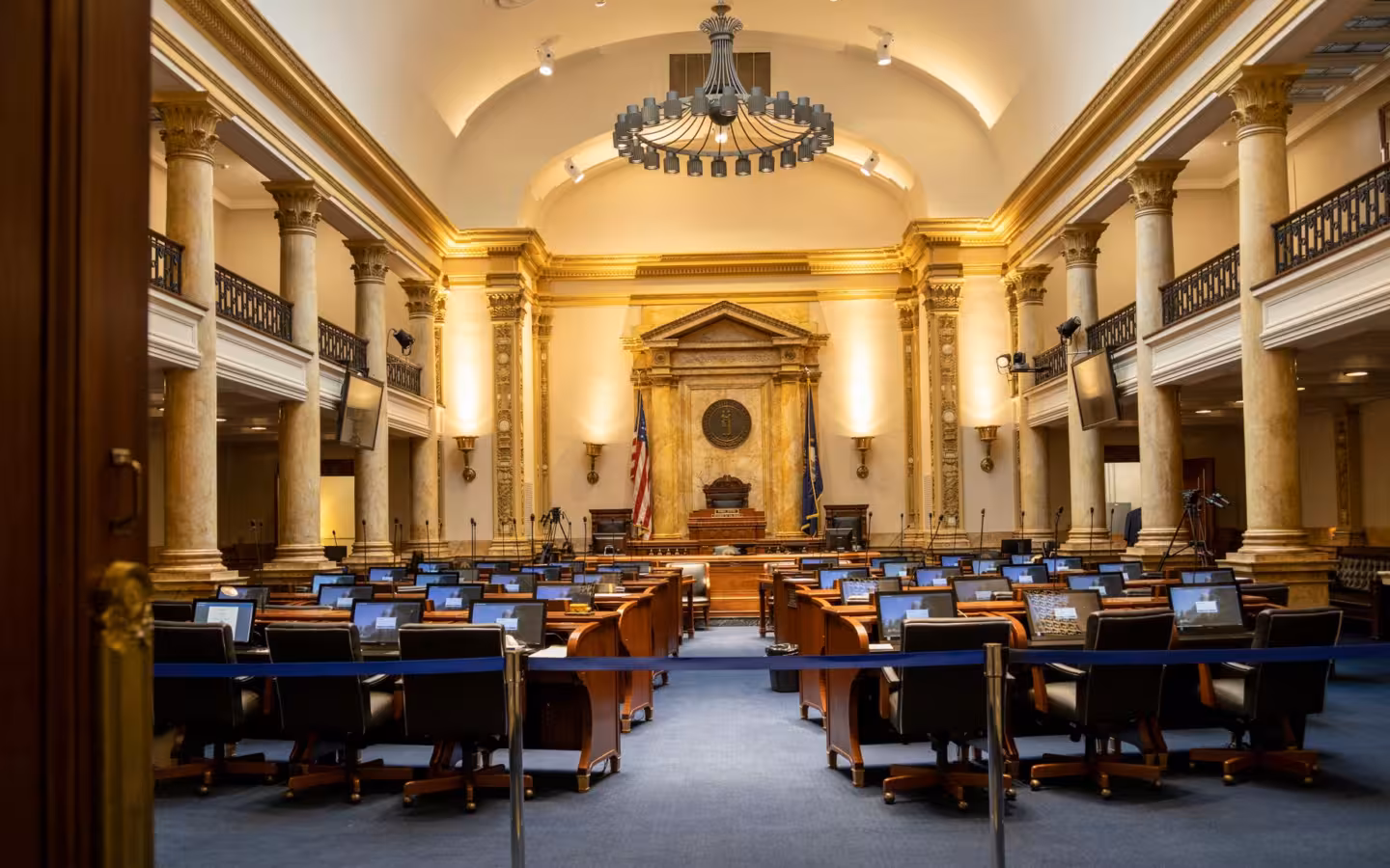 An empty government chamber with columns, desks, screens and a chandelier