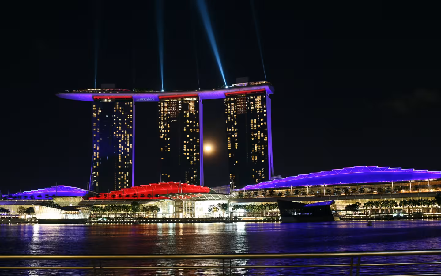 Marina Bay Sands illuminated at night, with light beams above the waterfront.