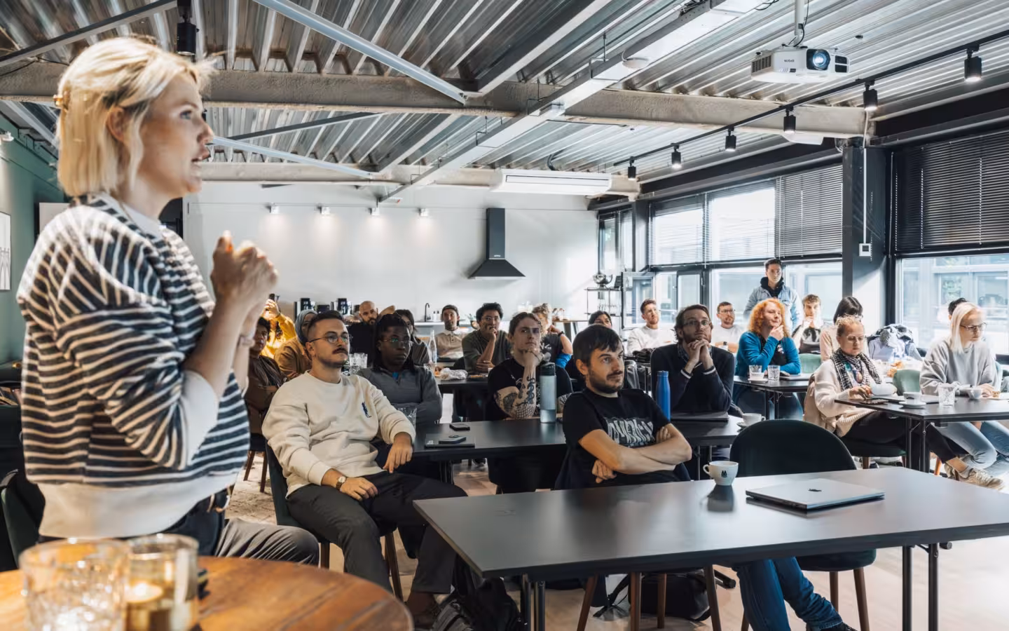 A speaker addresses an audience seated at tables in a modern workshop.