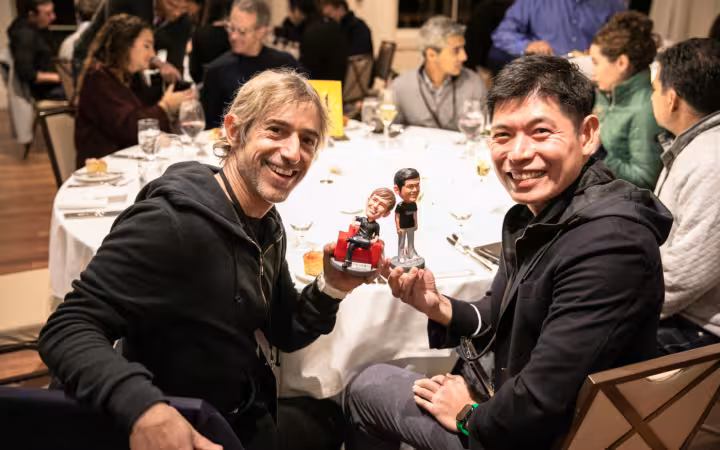 Mark Pincus and Anthony Tan smile at a dinner, holding figurines.