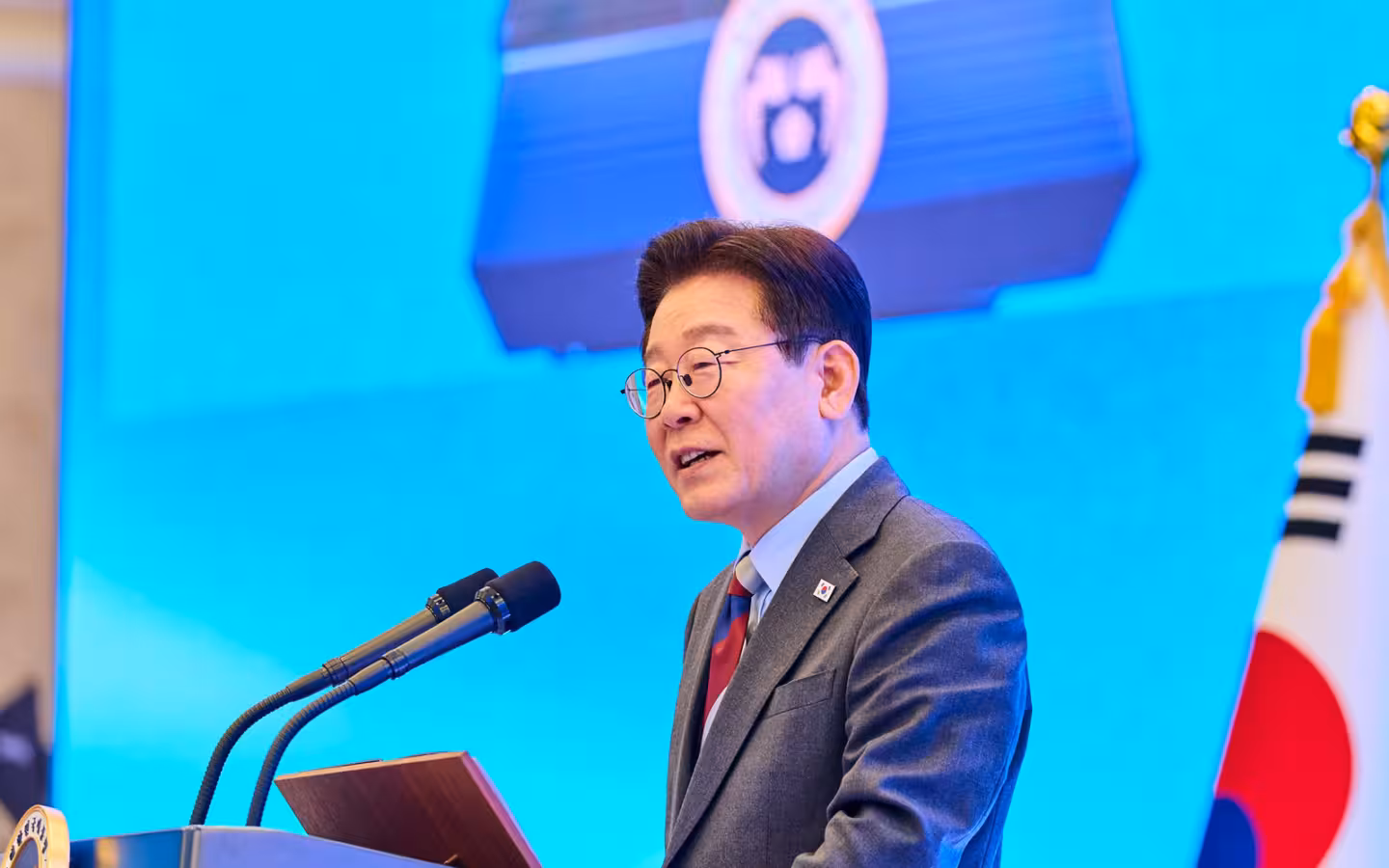 President Lee Jae Myung speaking at a lectern beside a South Korean flag