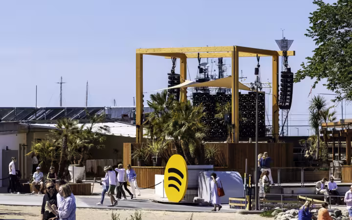Spotify’s yellow logo stands before an outdoor stage and scattered visitors.