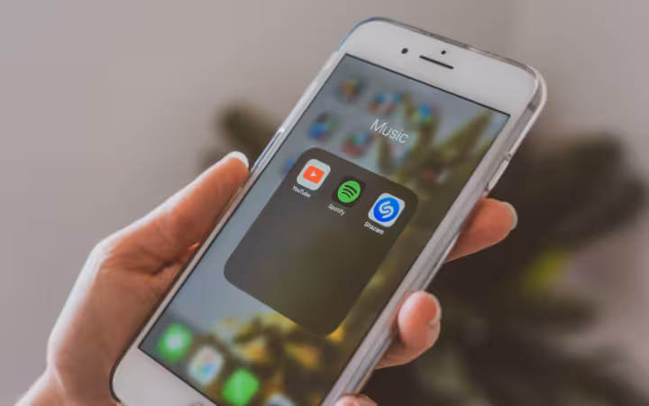 A hand holds a smartphone showing YouTube, Spotify and Shazam music apps.