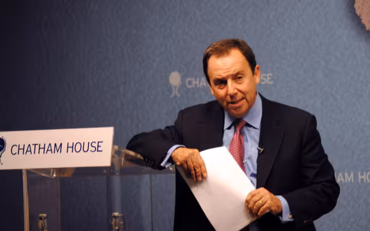 Ron Suskind stands beside a Chatham House lectern holding papers.