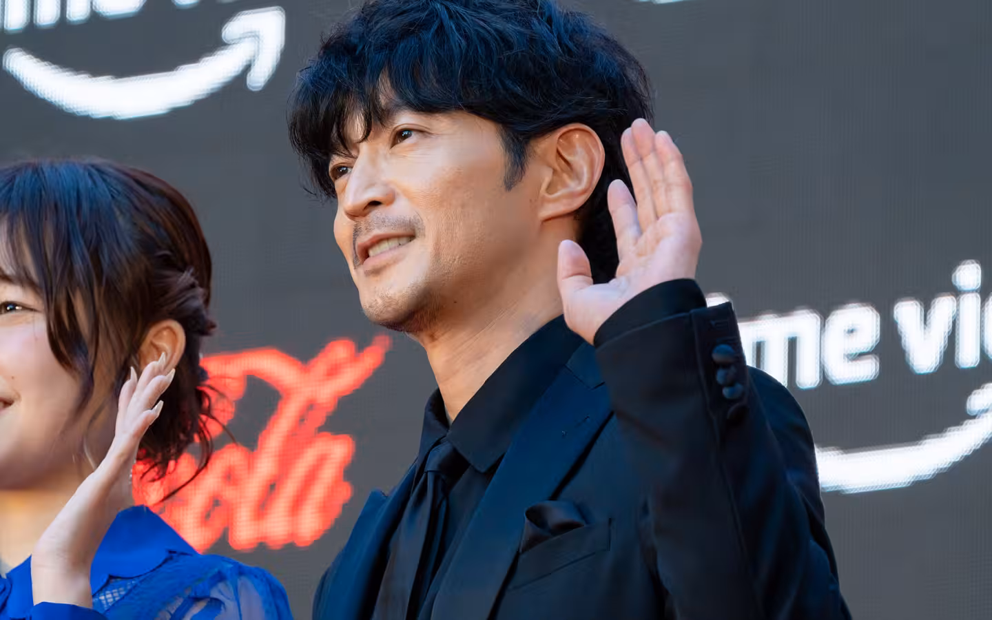 Kenjiro Tsuda waves beside a partially visible woman at a film festival.