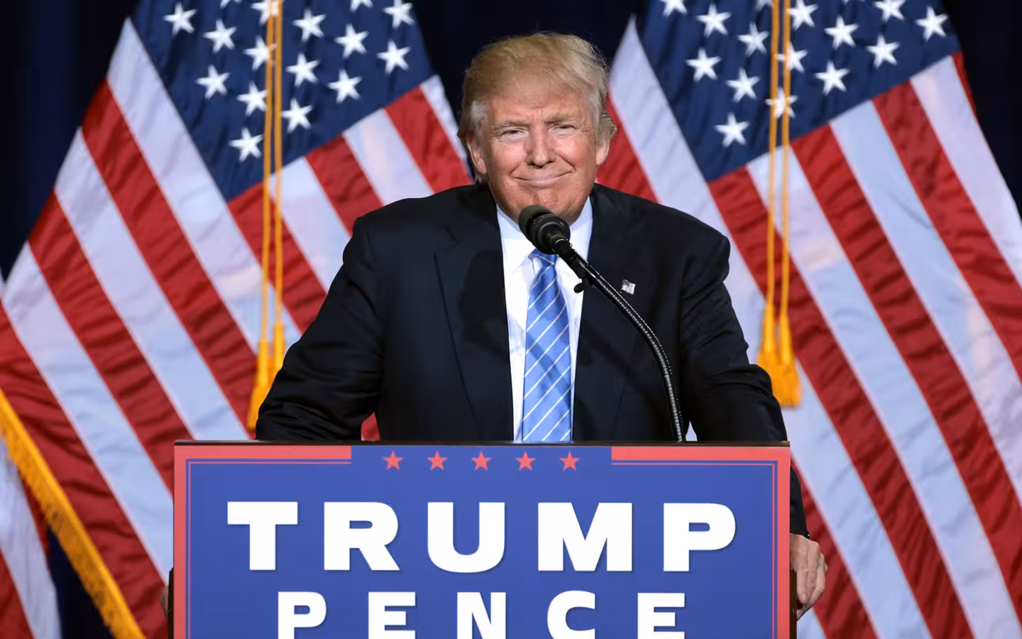 Donald Trump speaking behind a lectern with American flags behind him.
