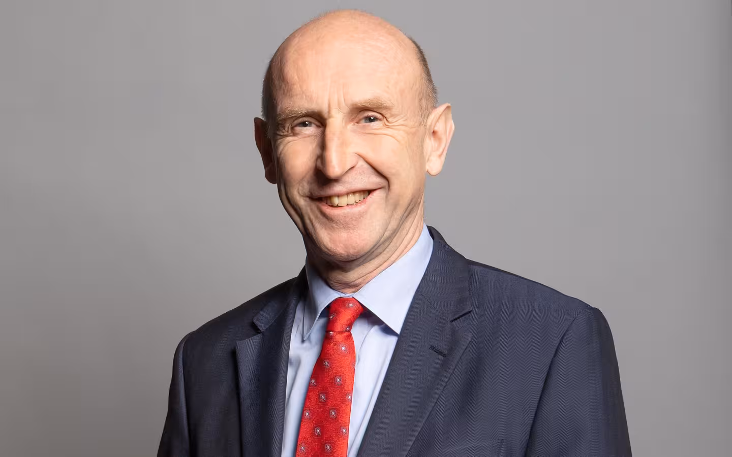 John Healey smiling in a suit and red tie against a grey background