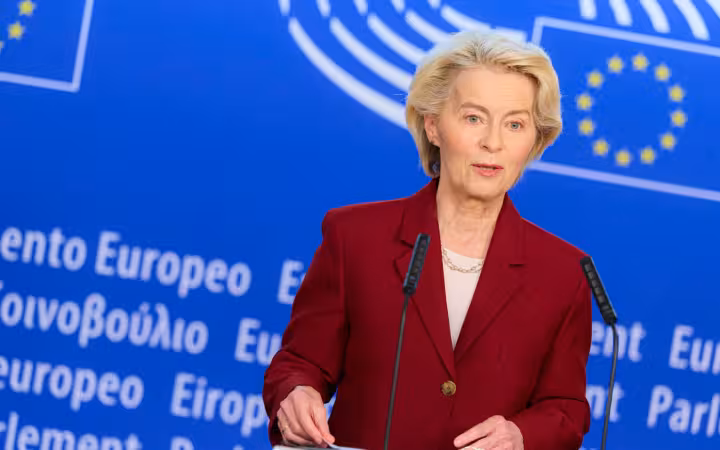 Ursula von der Leyen speaks between microphones before a blue European Parliament backdrop.